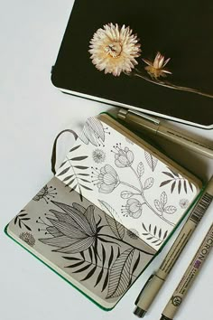 an open notebook with a flower on it and two markers next to it, along with a pen