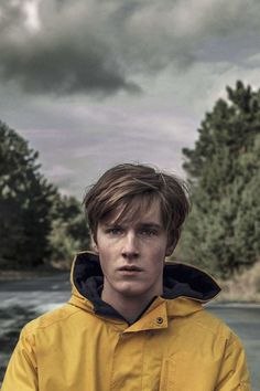 a young man in a yellow raincoat standing on the side of a river under a cloudy sky