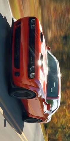 a red sports car driving down a road in the fall time with its lights on