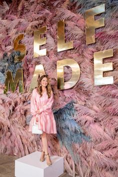 a woman standing in front of a pink wall with gold letters and feathers on it