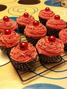 twelve cupcakes with red frosting on a cooling rack in front of a table