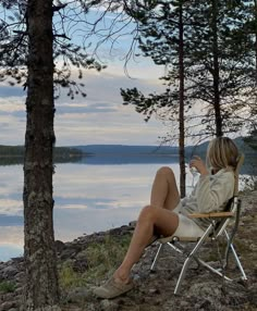 a woman sitting in a chair on the shore of a lake with her legs crossed