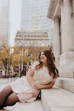 a woman sitting on the side of a stone wall in front of tall buildings with her legs crossed