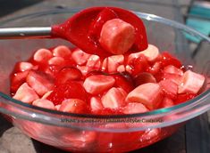 a glass bowl filled with sliced strawberries on top of a wooden table next to a metal spoon