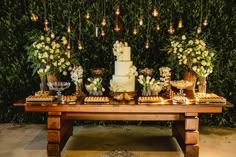 a table topped with lots of desserts and flowers next to a wall covered in greenery