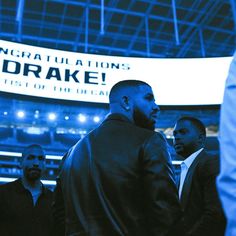 two men standing in front of a large sign at a sporting event, one is wearing a black jacket and the other wears a white shirt