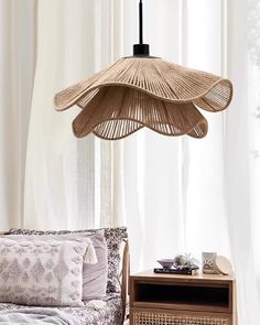 a wicker lamp hanging over a bed next to a night stand and table in front of a window