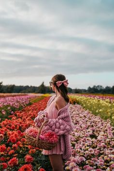 a woman standing in a field full of flowers holding a basket with pink and red flowers