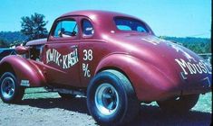 an old red truck with writing on it's side sitting in the dirt next to other cars