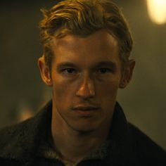 a man with blonde hair wearing a black jacket and looking at the camera while standing in a dark room