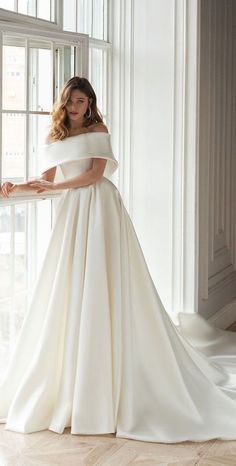 a woman standing in front of a window wearing a white wedding dress with off the shoulder sleeves