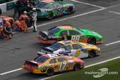 three cars racing on a race track with people watching from the sidelines behind them