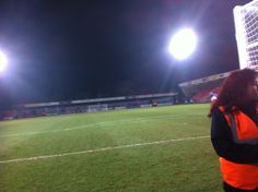 a woman in an orange vest standing on a soccer field at night with the lights on