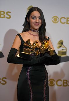 a woman in a black dress holding two golden trophies and posing for the camera with her hands on her hips