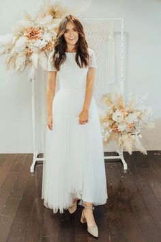 a woman wearing a white dress standing in front of a wall with flowers and feathers