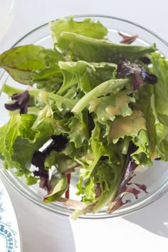 a glass bowl filled with lettuce on top of a white table next to a blue and white plate