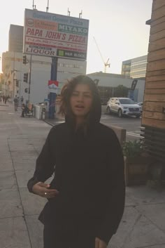 a woman standing on the sidewalk in front of a pizza place with cars and buildings behind her