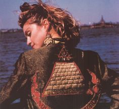 a woman standing in front of the ocean with her back to the camera, wearing a jacket and earrings