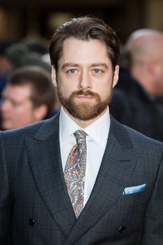 a man with a beard wearing a suit and tie in front of other people at an event