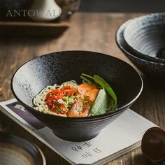 a black bowl filled with noodles and vegetables on top of a wooden table next to chopsticks