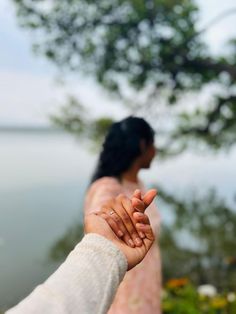 a woman holding the hand of another person near some flowers and water with trees in the background