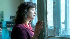 a woman standing in front of a window holding her hands up and looking out the window