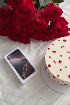 a white cake sitting on top of a table next to red roses and a cell phone