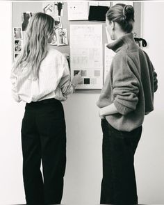 two women standing next to each other in front of a wall with papers on it