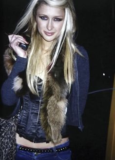 a woman with long blonde hair wearing a fur vest and holding a cell phone to her ear