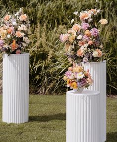 three white vases with flowers in them sitting on the grass next to some bushes