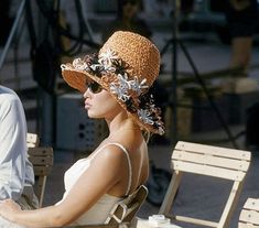 a woman sitting on a wooden bench wearing a hat with flowers in it's brim