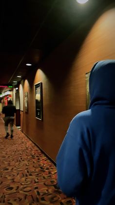 two people walking down a long hallway in a hotel or office building, one is wearing a blue jacket and the other has a black hoodie