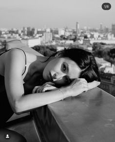 a black and white photo of a woman leaning her head on a ledge with the city in the background