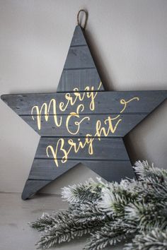 a wooden star hanging on the wall with merry and bright written in gold ink next to evergreen branches