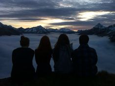 three people sitting on top of a hill looking at the sky with clouds and mountains in the background