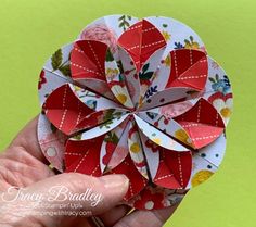 a hand holding up a paper flower with red and white flowers on it, in front of a green background