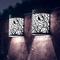 two lamps that are sitting on the side of a stone wall at night with trees and birds in them