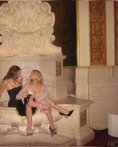 two women sitting on a bench in front of a fountain with wine glasses and champagne