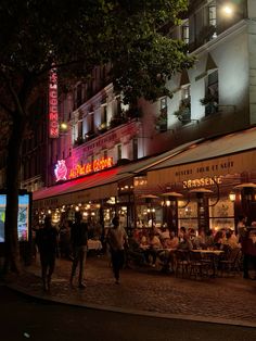 people are sitting at tables on the sidewalk in front of a building with neon lights