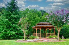 a gazebo in the middle of a park surrounded by trees and bushes with purple flowers