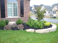 a brick house with green grass and bushes in the front yard, next to a large rock garden bed