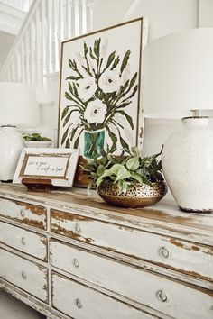 a white dresser with flowers and framed pictures on it's sideboard next to a stair case