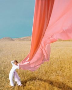 a woman standing in the middle of a field holding onto a pink cloth over her head