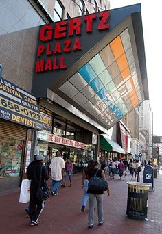 people walking on the sidewalk in front of a pizza mall with a large sign that says gertz plaza mall