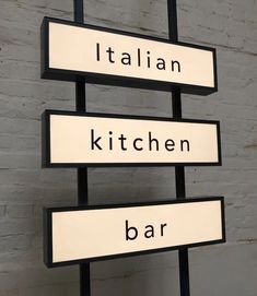 three black and white street signs with italian, kitchen, bar written on them against a brick wall