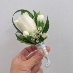 a hand holding a white flower with green leaves and pearls on the end, in front of a gray background