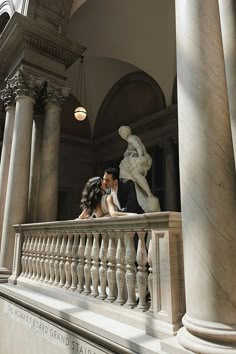 a man and woman kissing on the balcony of a building with statues in the background