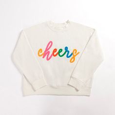 A cute, cozy, and colorful sweater is a must-have for your winter wardrobe! Stay warm throughout the Christmas season and into the New Year with our Large Cheers Sweatshirt! Chest measures 24.8 inches Sleeve length measures 33.8 inches Crafted of 84.6% cotton and 15.4% polyester Features the text "Cheers" Accented with soft, embroidered fabric Hues of white, yellow, pink, orange, green, and blue Care: Machine wash cold. Line dry. Contact your local Kirkland's store for availability. Quantities a Holiday Sweatshirts, Sweatshirt White, Holiday Sweatshirt, Embroidered Fabric, Seasonal Gifts, Holiday Shirts, Green And Blue, Christmas Season, Christmas Seasons