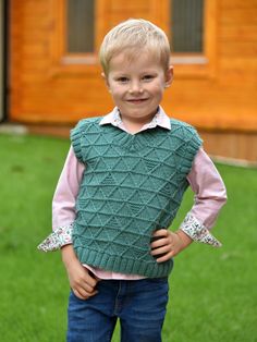 a young boy wearing a green sweater vest and jeans standing in front of a wooden building