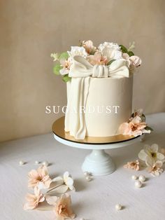 a white cake with pink flowers on top is sitting on a table and surrounded by petals